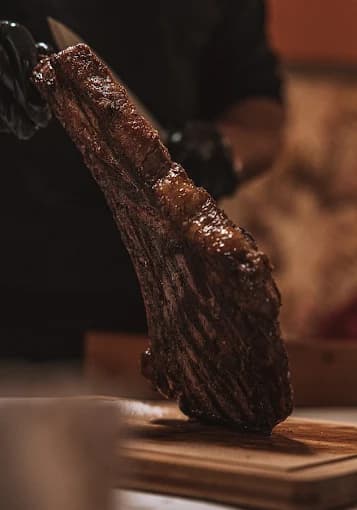 A large, seared steak held vertically over a wooden cutting board by gloved hands.