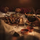 People sharing a meal of grilled meat, wine, and side dishes under warm, dim lighting.