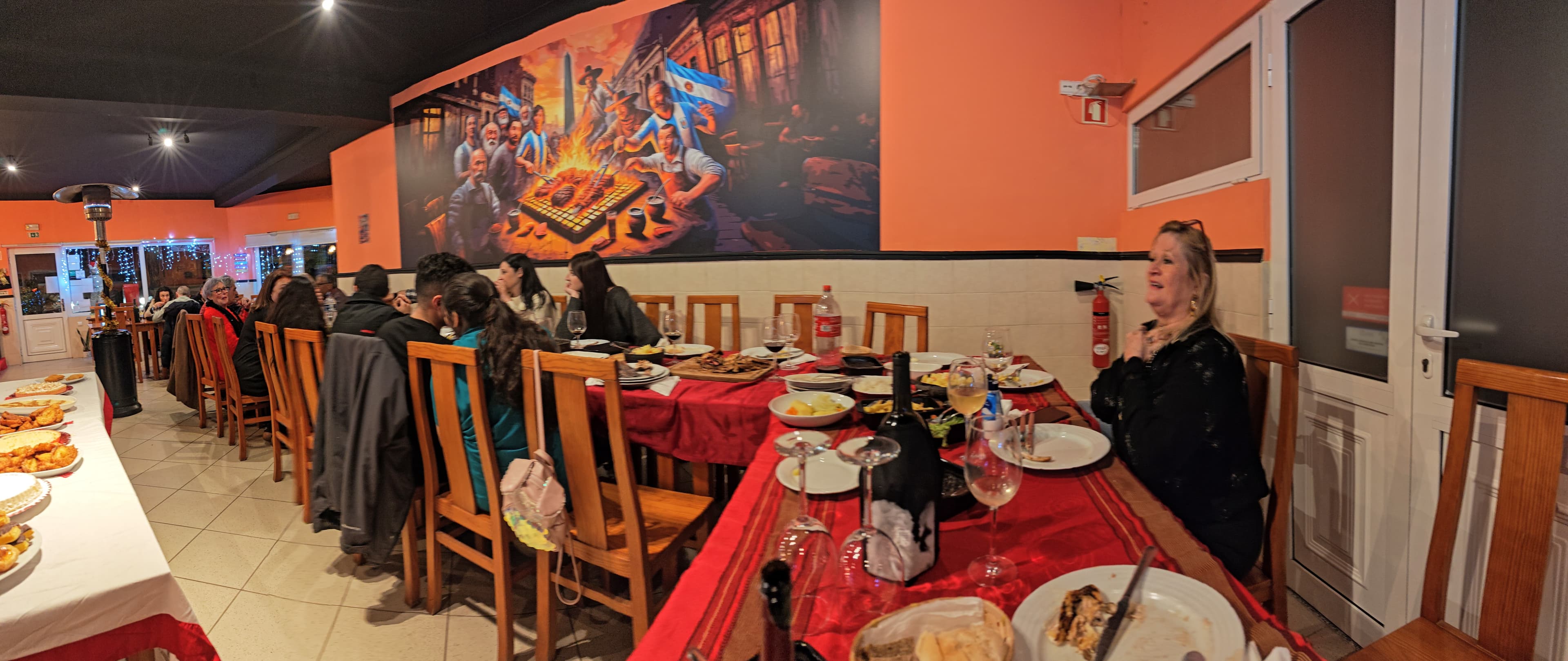 Diners at long tables in a restaurant with orange walls and a large Argentinian-themed mural.